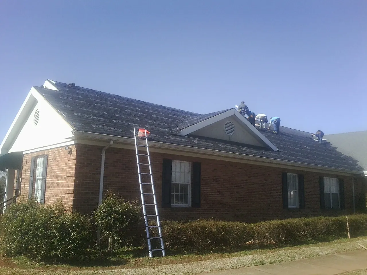 Crew tearing shingles off a brick ranch roof with a ladder set — Metal Roof Repair work by our crew in Vernon
